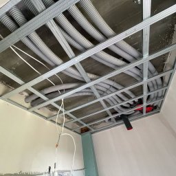ENKAM SPÓŁKA Z OGRANICZONĄ ODPOWIEDZIALNOŚCIĄ - Szare rury wentylacyjne poprowadzone pod sufitem podwieszanym, widoczne elementy instalacji elektrycznej. Kątowe ujęcie z dołu, surowy stan wykończenia.