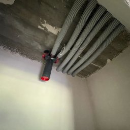 ENKAM SPÓŁKA Z OGRANICZONĄ ODPOWIEDZIALNOŚCIĄ - Szare, karbowane rury wentylacyjne zamontowane pod surowym betonowym sufitem, z czarnym rozdzielaczem z czerwonymi elementami. Widok z dołu w rogu pomieszczenia.