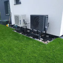 Zewnętrzna jednostka pompy ciepła AUX, zamontowana na metalowej konstrukcji przy białej ścianie domu. Widoczny fragment trawnika i kostki brukowej.