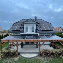 Dom z panelami słonecznymi na dachu i nad tarasem. Na dachu czarna dachówka, na tarasie panele na drewnianej konstrukcji. Pochmurne niebo w tle.