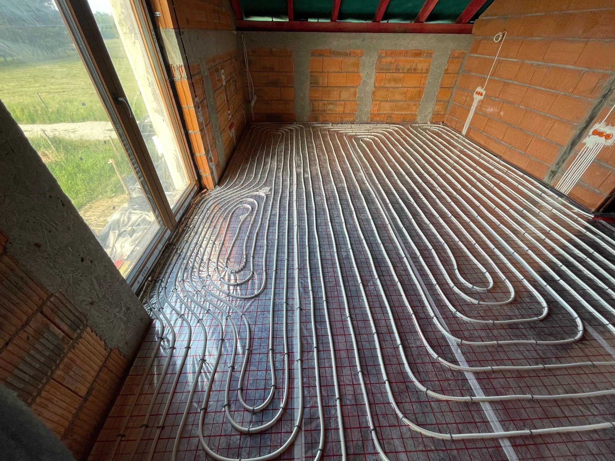 Instalacja ogrzewania podłogowego w surowym wnętrzu. Białe rury ułożone na folii z siatką, widoczne ceglane ściany i okno z widokiem na zewnątrz.