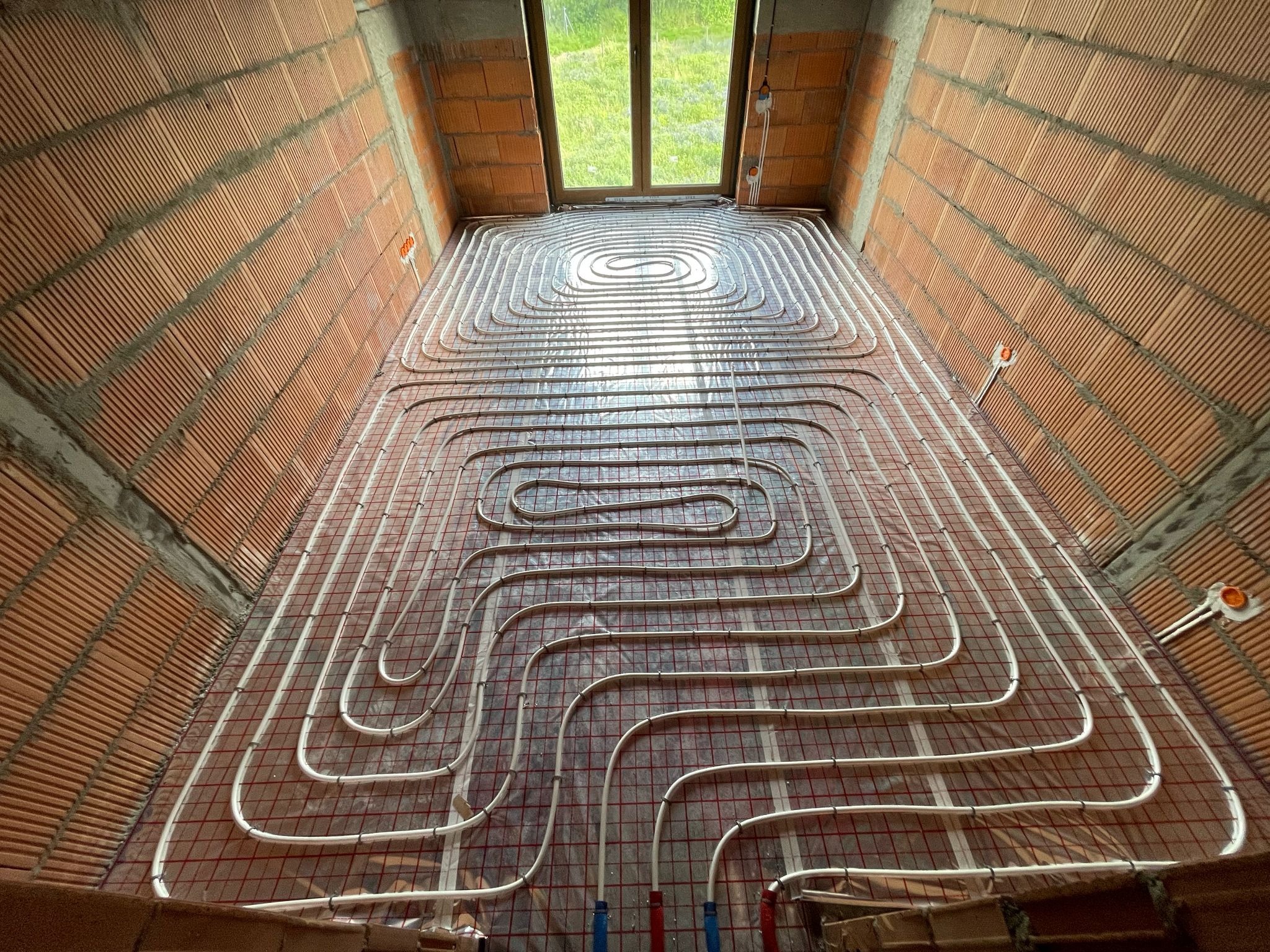Instalacja ogrzewania podłogowego w surowym wnętrzu z ceglanymi ścianami. Białe rurki ułożone spiralnie na folii z siatką. Widok z góry na okno.