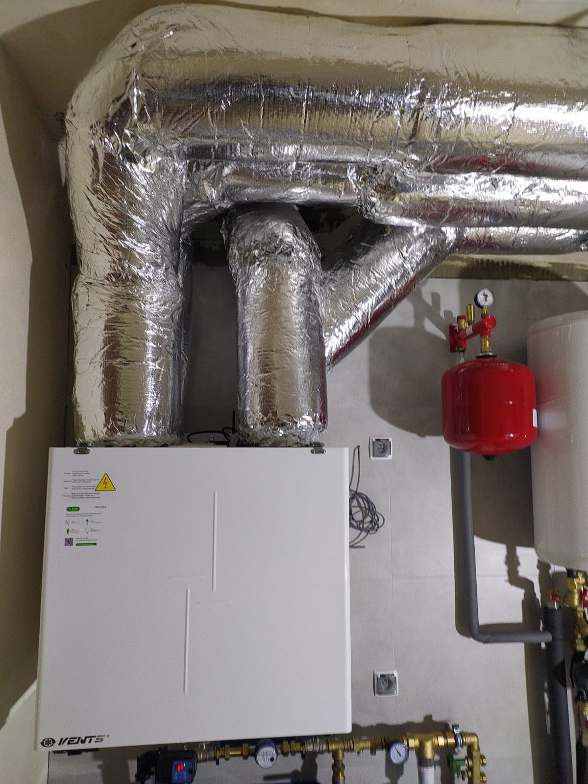 Biała centrala wentylacyjna Vents z izolowanymi, srebrnymi przewodami wentylacyjnymi, czerwonym zbiornikiem i osprzętem hydraulicznym na tle szarej ściany.