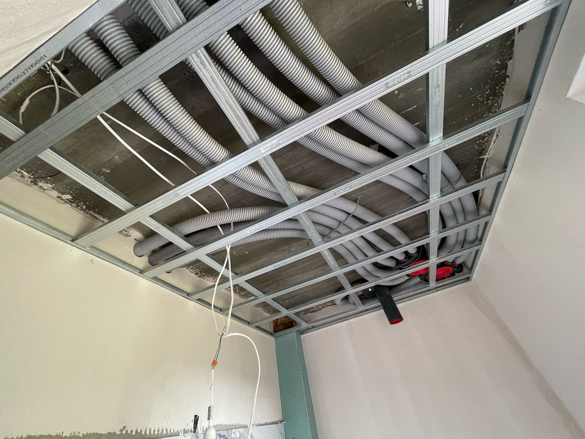 Szare rury wentylacyjne poprowadzone pod sufitem podwieszanym, widoczne elementy instalacji elektrycznej. Kątowe ujęcie z dołu, surowy stan wykończenia.