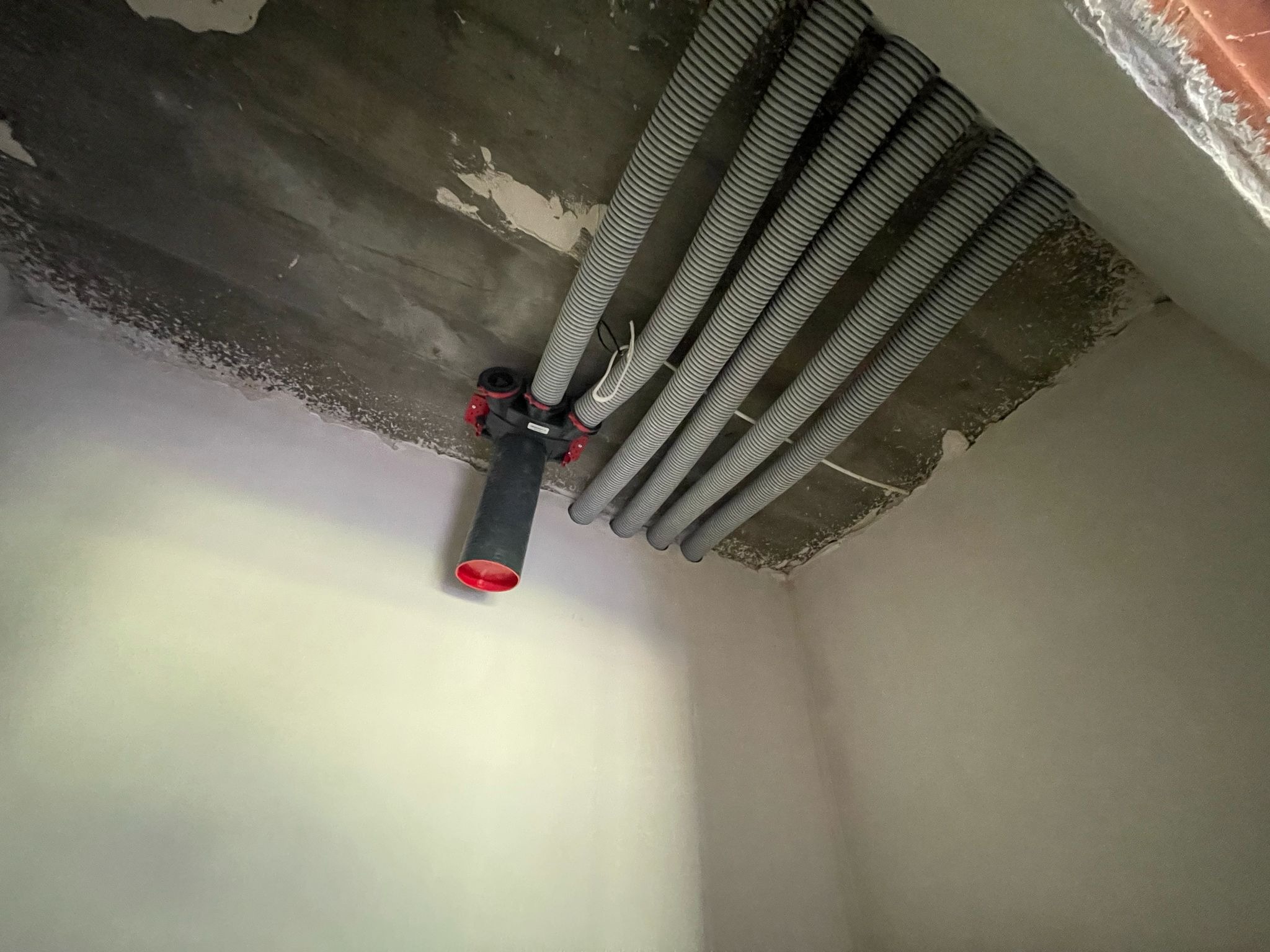 Szare, karbowane rury wentylacyjne zamontowane pod surowym betonowym sufitem, z czarnym rozdzielaczem z czerwonymi elementami. Widok z dołu w rogu pomieszczenia.