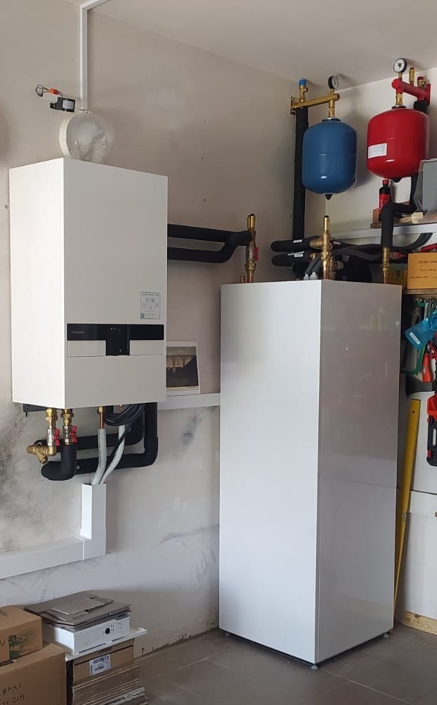 Biały, nowoczesny piec grzewczy z podłączonymi rurami, zbiornik wyrównawczy (czerwony i niebieski) na ścianie w pomieszczeniu gospodarczym. Widoczne narzędzia.