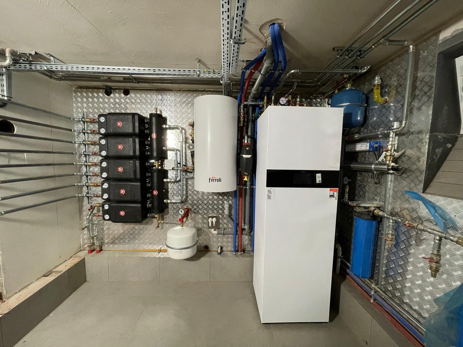 Kotłownia z piecem Ferroli, zbiornikiem i armaturą. Instalacja grzewcza z licznymi rurami miedzianymi i elementami zabezpieczającymi na ścianie wyłożonej blachą aluminiową.