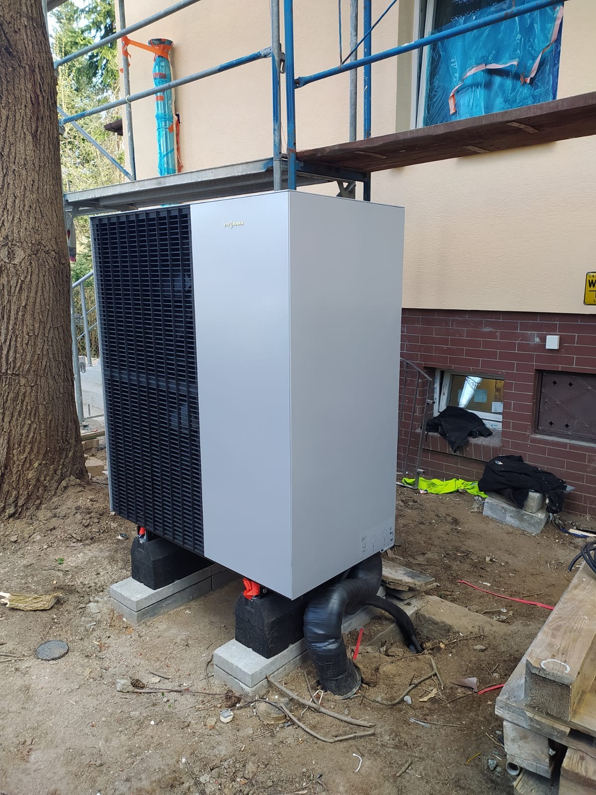 Szara pompa ciepła Viessmann ustawiona na betonowych blokach przy budynku. Widoczne rury i rusztowanie, sugerujące etap instalacji.