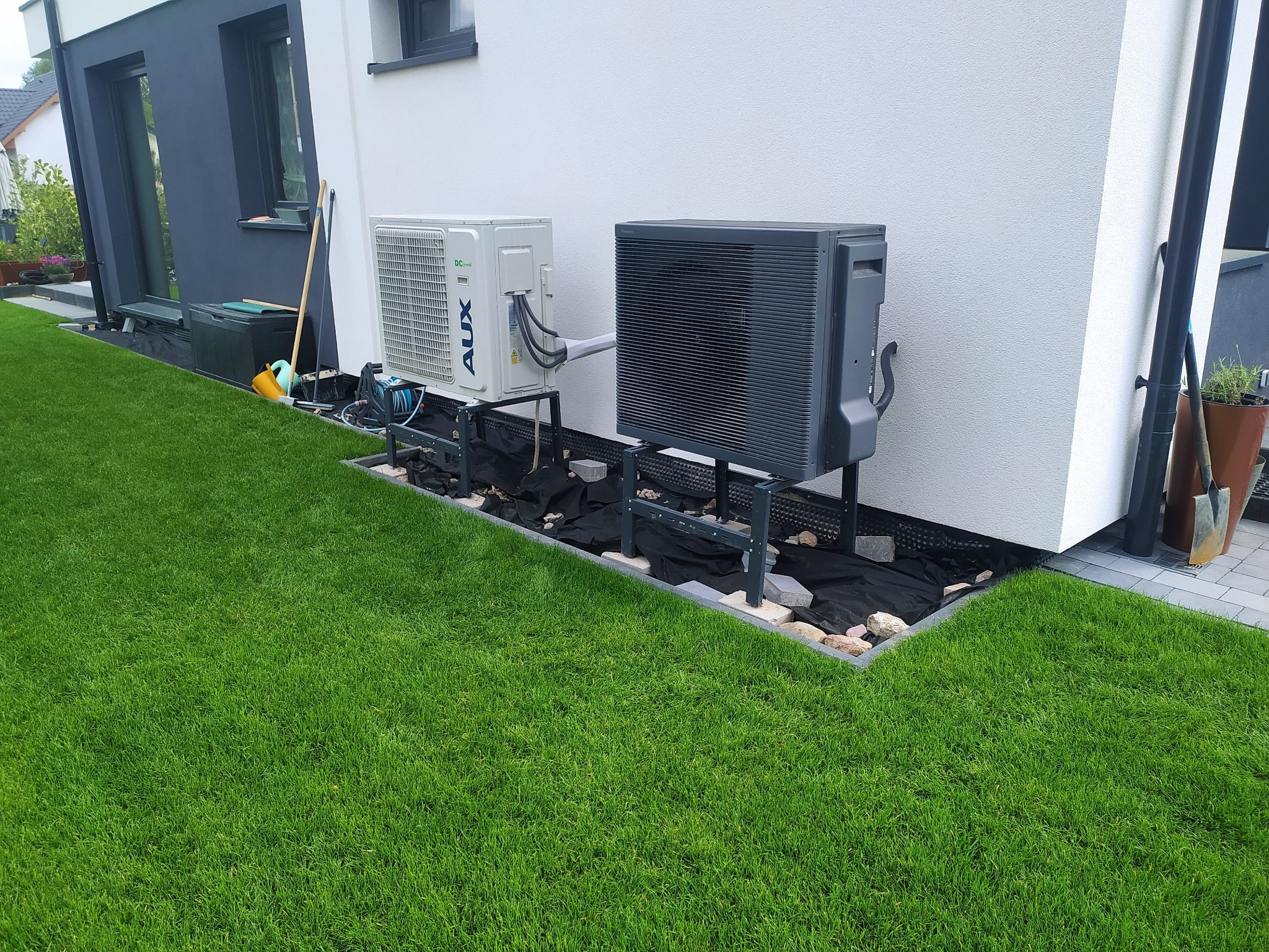Zewnętrzna jednostka pompy ciepła AUX, zamontowana na metalowej konstrukcji przy białej ścianie domu. Widoczny fragment trawnika i kostki brukowej.