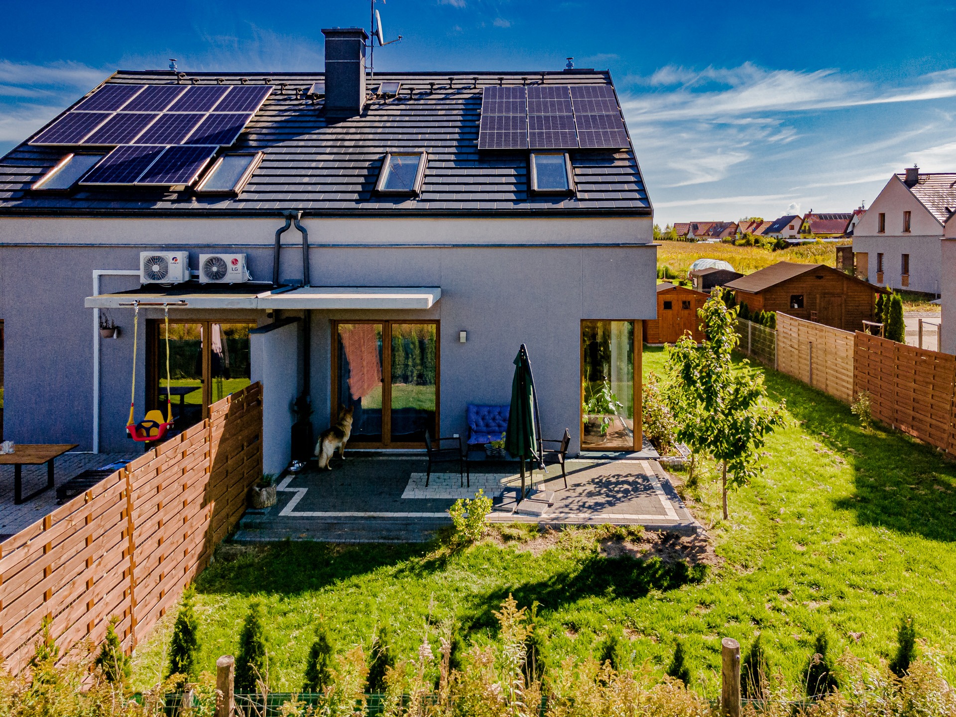 Dom jednorodzinny z panelami fotowoltaicznymi na dachu i klimatyzacją na ścianie. Widoczny ogród i pies przy wejściu.