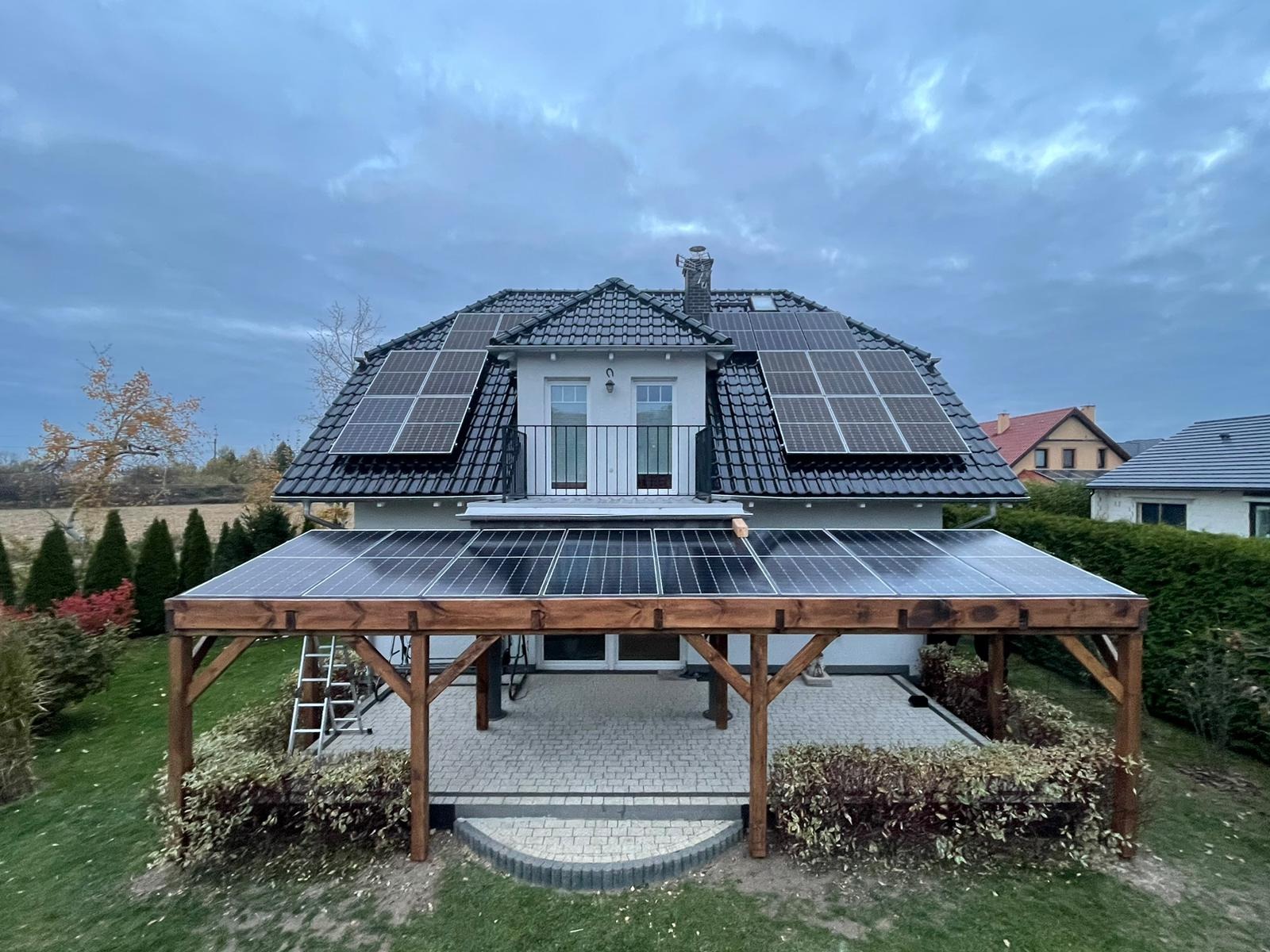 Dom z panelami słonecznymi na dachu i nad tarasem. Na dachu czarna dachówka, na tarasie panele na drewnianej konstrukcji. Pochmurne niebo w tle.