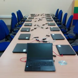 Sala konferencyjna z długim stołem, na którym rozstawione są laptopy z podłączonymi zasilaczami i kablami sieciowymi, widoczna płyta CD, niebieskie krzesła biurowe.