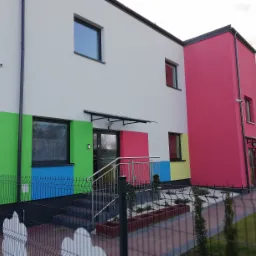 Wielobarwny budynek z domofonem przy wejściu, widoczny zza ogrodzenia w Zduńskiej Woli.