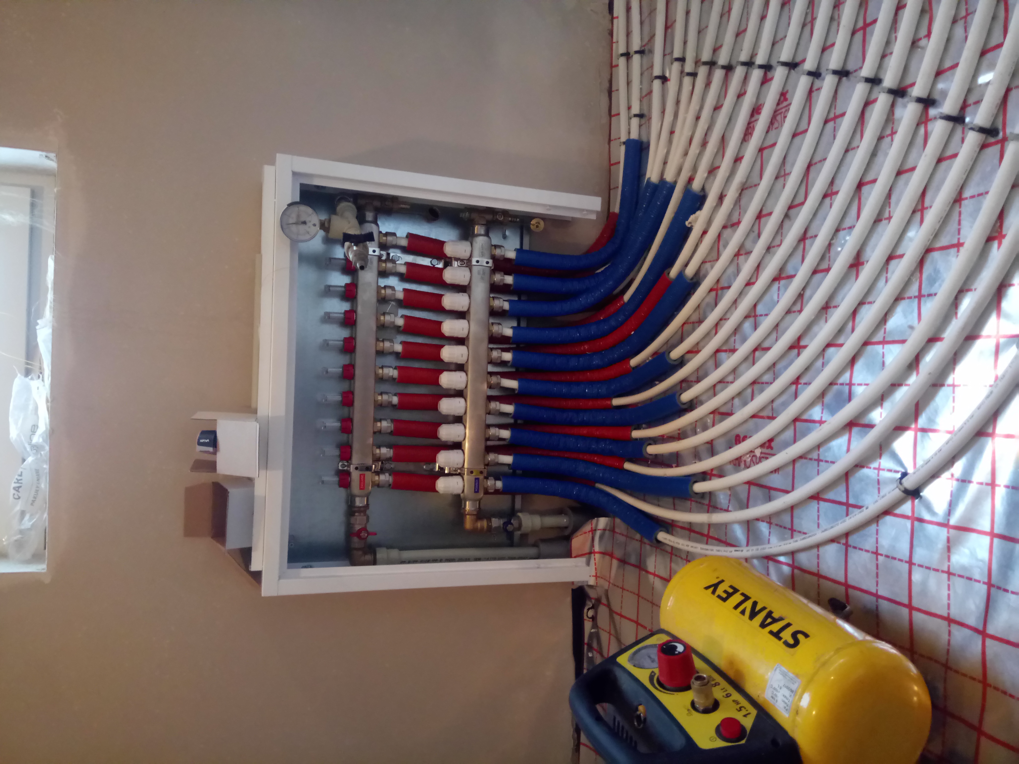Rozdzielacz ogrzewania podłogowego w białej szafce, z rurami PEX oznaczonymi kolorami, oraz żółty kompresor Stanley w tle na ścianie z izolacją.