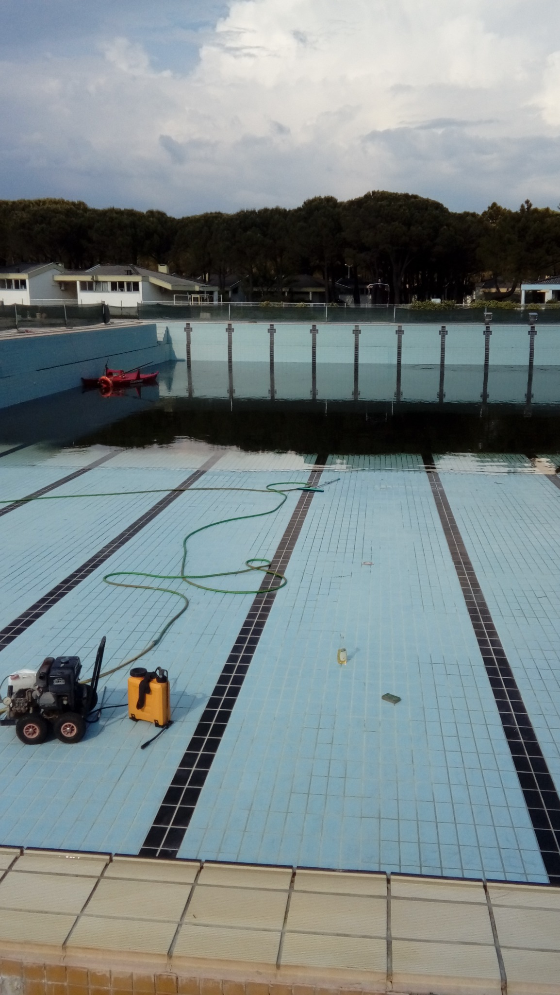 Opróżniony basen z resztkami wody i glonów, sprzęt czyszczący na kafelkach, zielony wąż, w tle budynek i drzewa pod pochmurnym niebem.