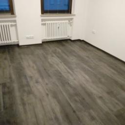 Jacek Kuchnicki - Szara podłoga z paneli imitujących drewno, ułożona w pustym pokoju z białymi ścianami i grzejnikami pod oknami.