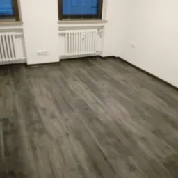 Szara podłoga z paneli imitujących drewno, ułożona w pustym pokoju z białymi ścianami i grzejnikami pod oknami.