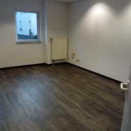 Jacek Kuchnicki - Pusty pokój po remoncie z panelami podłogowymi w odcieniach brązu, białymi ścianami, grzejnikiem i widokiem z okna na białe ogrodzenie.