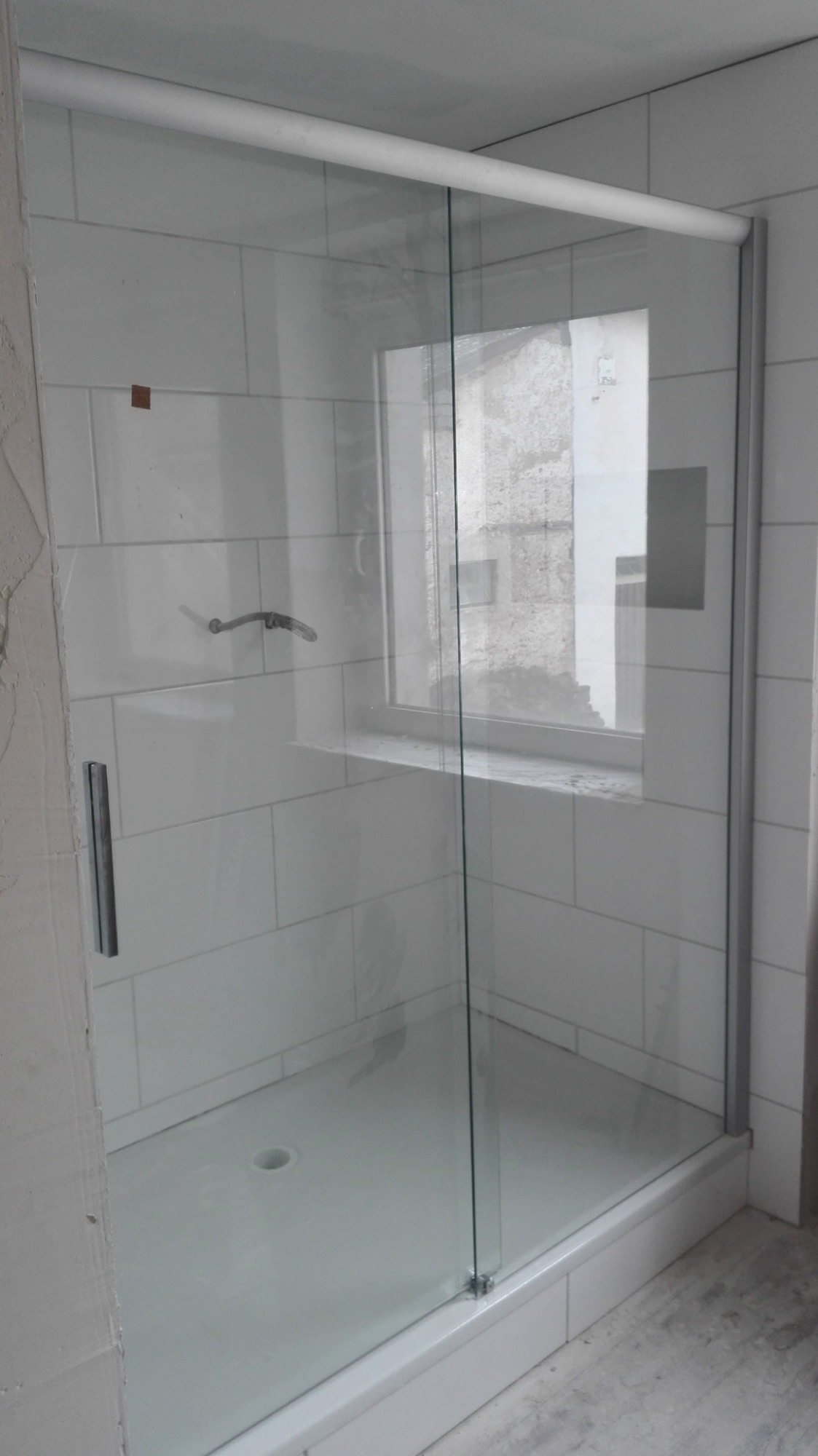 Nowoczesna kabina prysznicowa ze szklanymi drzwiami i białymi płytkami na ścianach, widoczny fragment niedokończonej ściany z surowym tynkiem.