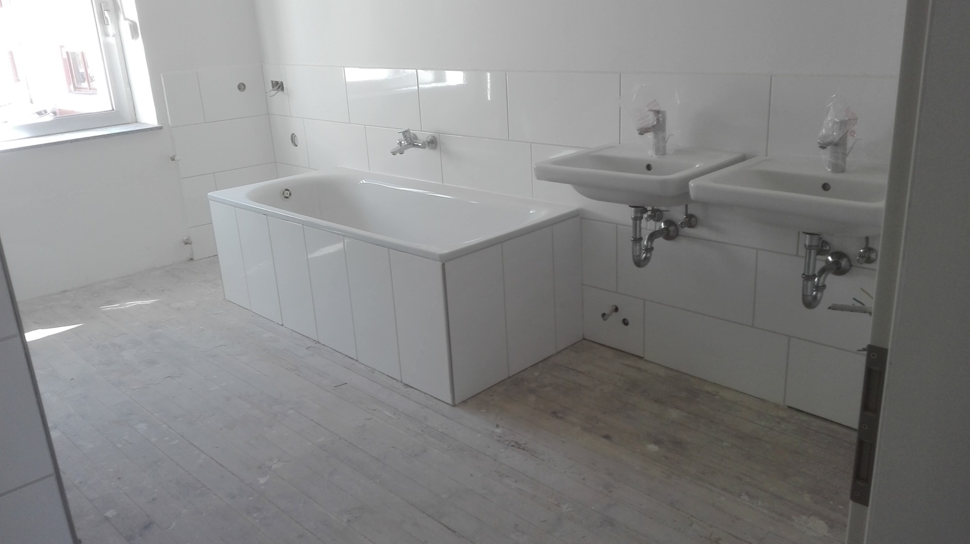 Nowa, jasna łazienka z białą wanną obudowaną panelami, dwiema umywalkami z bateriami w folii ochronnej i białymi płytkami na ścianach, na podłodze surowy beton.