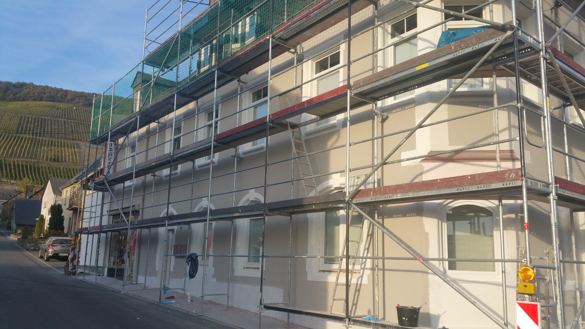 Elewacja domu w trakcie szpachlowania, widoczne rusztowania, częściowo nałożona szpachla, w tle winnice na wzgórzu.