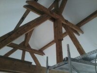 Wnętrze poddasza w trakcie prac wykończeniowych. Widoczne drewniane belki konstrukcyjne dachu na tle białej powierzchni sufitu oraz fragment rusztowania.