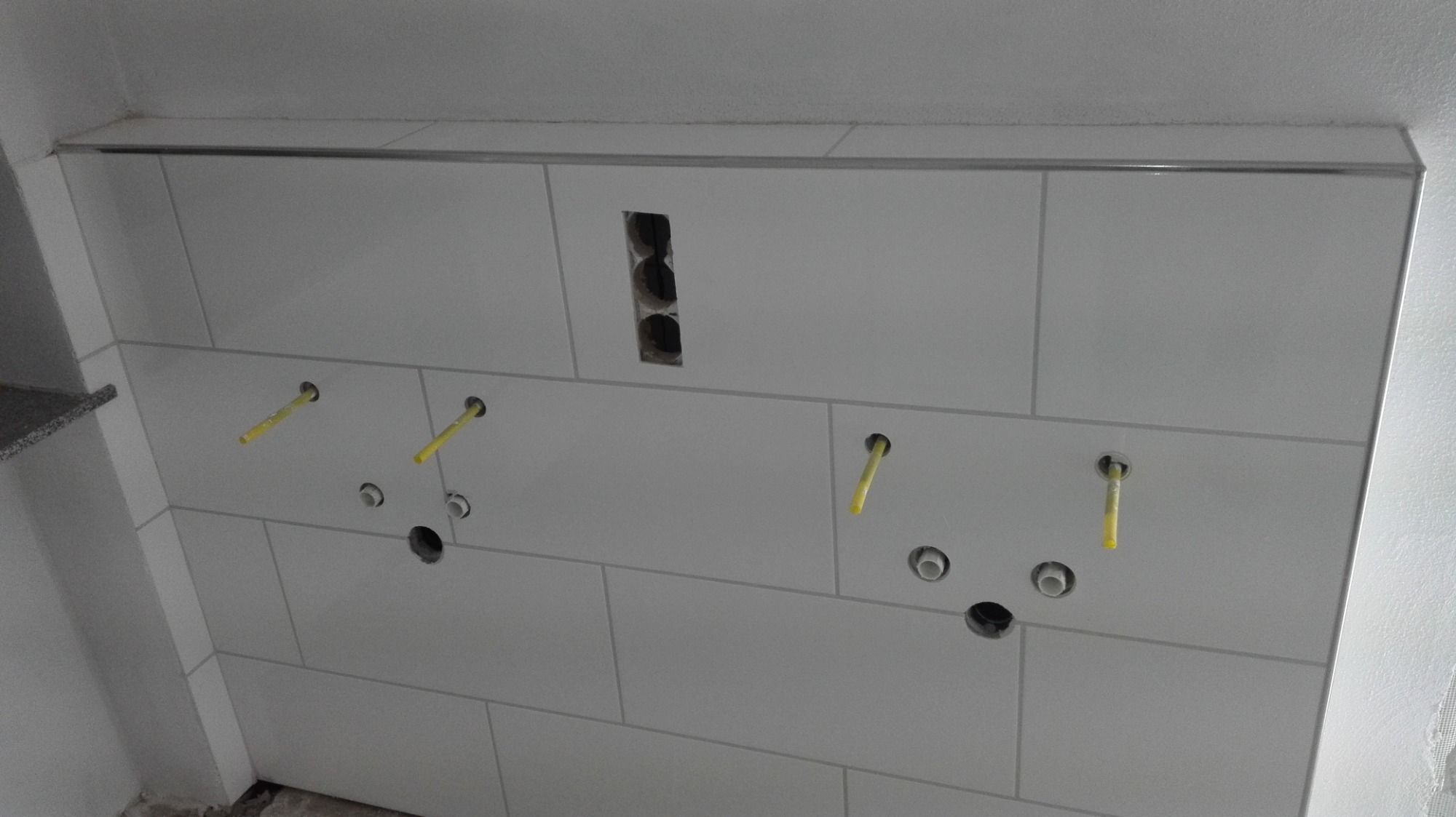 Ściana wyłożona białymi, prostokątnymi płytkami ceramicznymi z widocznymi otworami na instalacje wodno-kanalizacyjne i elektryczne, częściowo wykończona metalową listwą przy suficie.