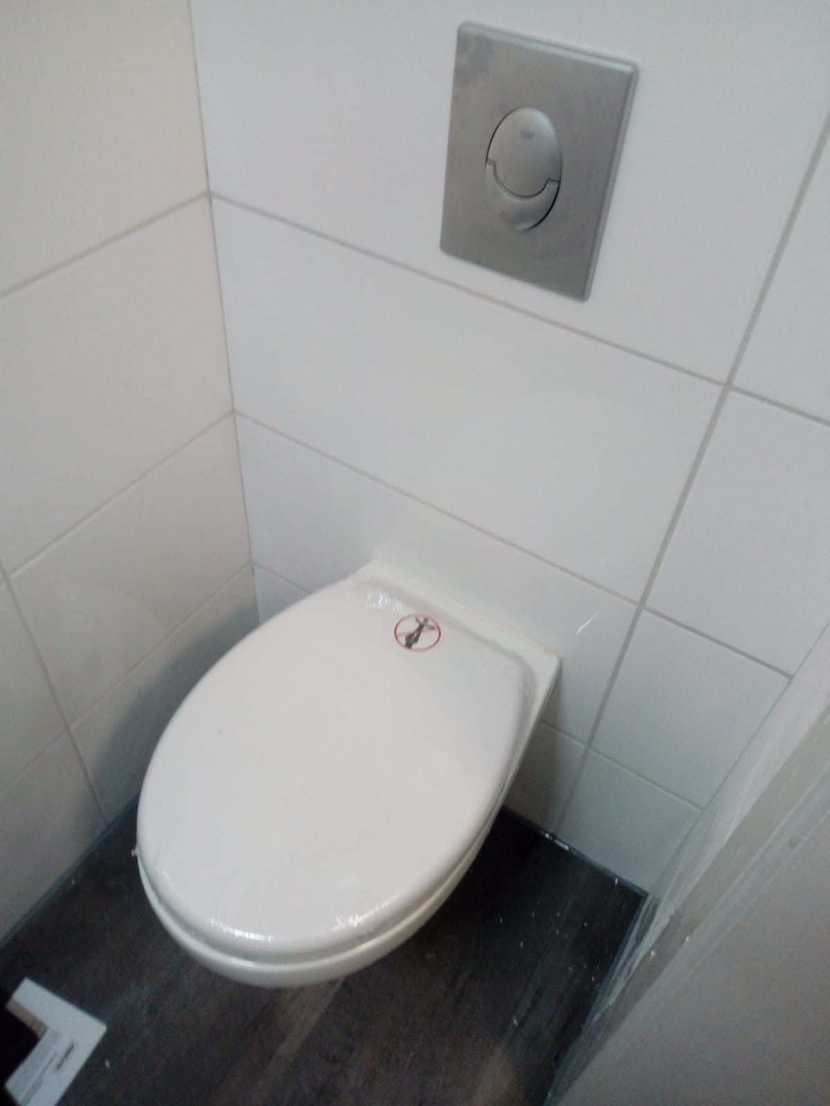 Nowa, biała toaleta wisząca z przyciskiem spłukującym na ścianie, zabezpieczona folią ochronną, ułożona na ciemnej podłodze imitującej drewno i otoczona białymi płytkami.