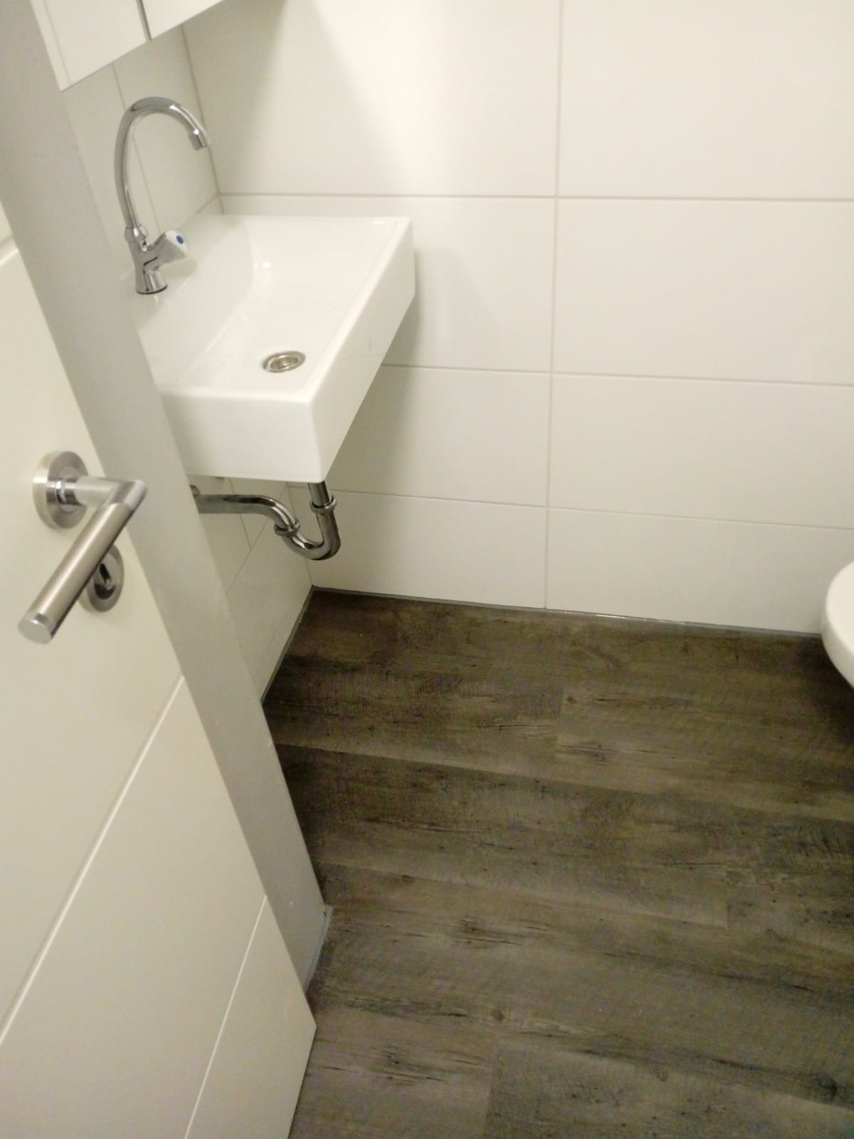 Nowoczesna, biała umywalka zamontowana na ścianie z chromowaną baterią i syfonem, widoczna na tle białych, kwadratowych płytek i podłogi z paneli imitujących drewno, z fragmentem otwartych, białych...