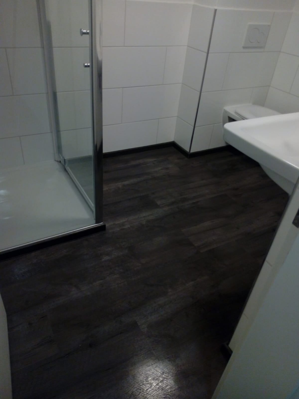 Wykończona łazienka z ciemną podłogą imitującą drewno, białe płytki na ścianach, kabina prysznicowa ze srebrnymi ramami i umywalka z toaletą w tle.