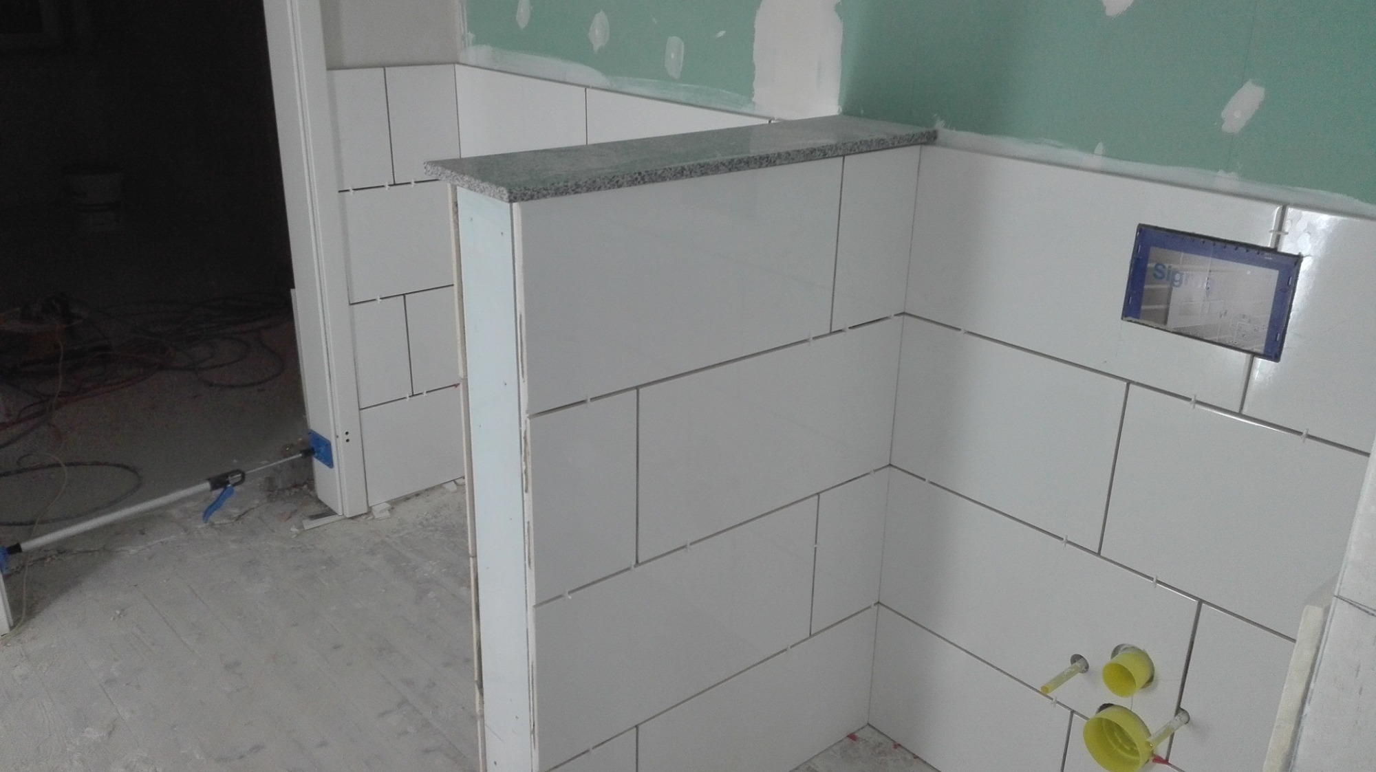 Wnętrze w trakcie wykańczania z białymi, prostokątnymi płytkami ceramicznymi na ścianach i blatem z szarego granitu, widoczne otwory na rury i puszka instalacyjna.