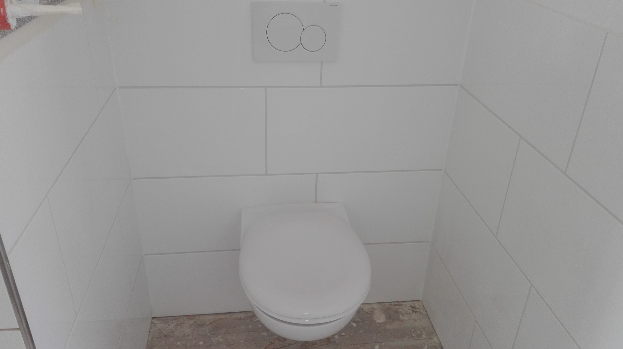 Biała wisząca toaleta w minimalistycznej łazience z białymi, prostokątnymi płytkami ułożonymi poziomo, z widocznym przyciskiem spłukującym Geberit na ścianie.