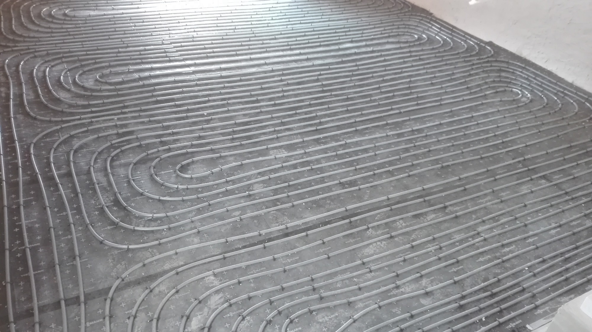 Szare rury ogrzewania podłogowego ułożone w pętle na szarym podłożu z widocznymi mocowaniami i oznaczeniami producenta.