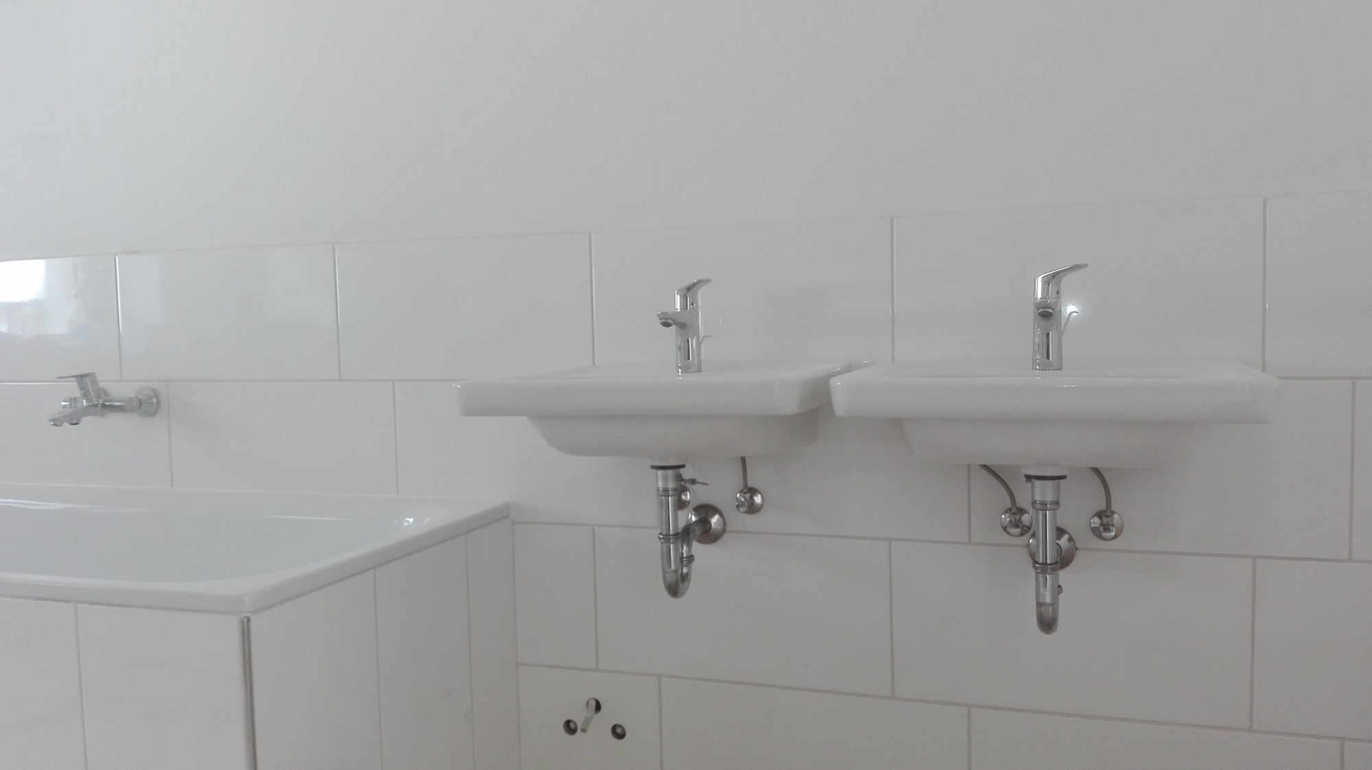 Biała łazienka z dwiema umywalkami i wanną wykończona białymi, prostokątnymi płytkami. Widoczne chromowane baterie i syfony. Na ścianie dodatkowy kran nad wanną.