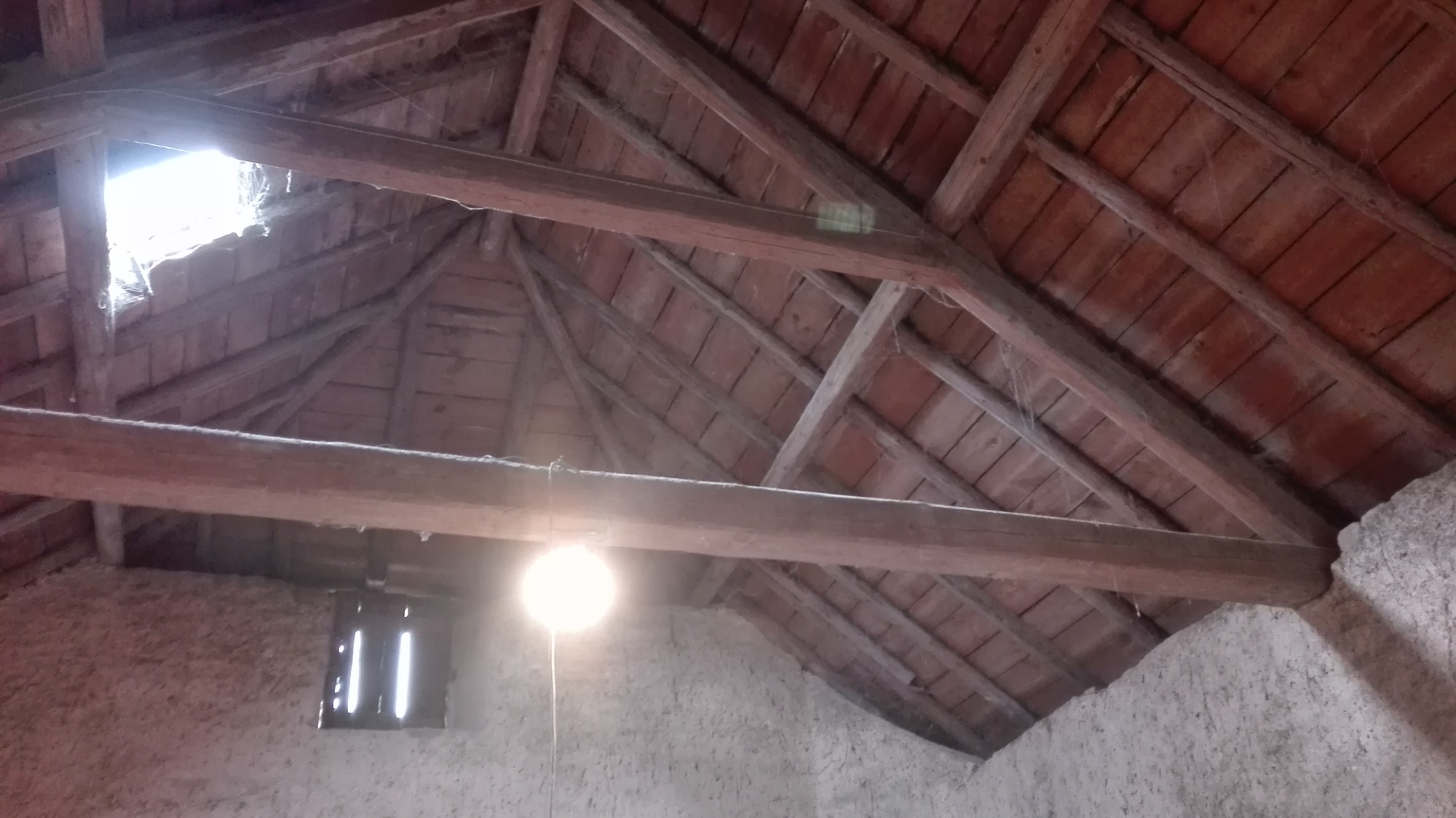 Wnętrze poddasza z drewnianą konstrukcją dachu, widoczne belki, deski i małe okienko, oświetlone przez wiszącą żarówkę i wpadające światło z góry, ściany o szorstkiej fakturze.