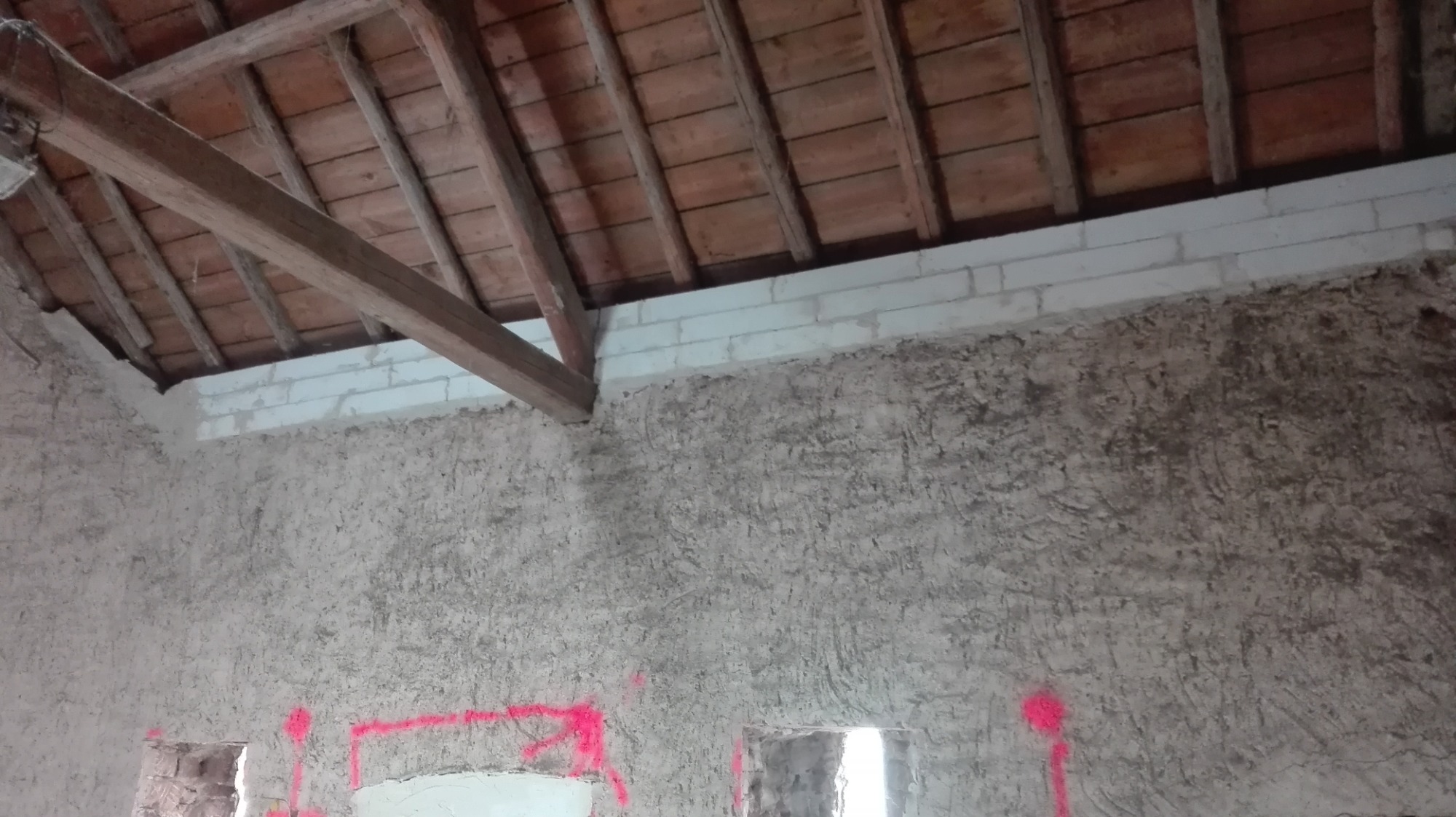 Wnętrze w trakcie renowacji: surowa ściana z ceglaną górną krawędzią, drewniany strop i różowe oznaczenia na ścianie.