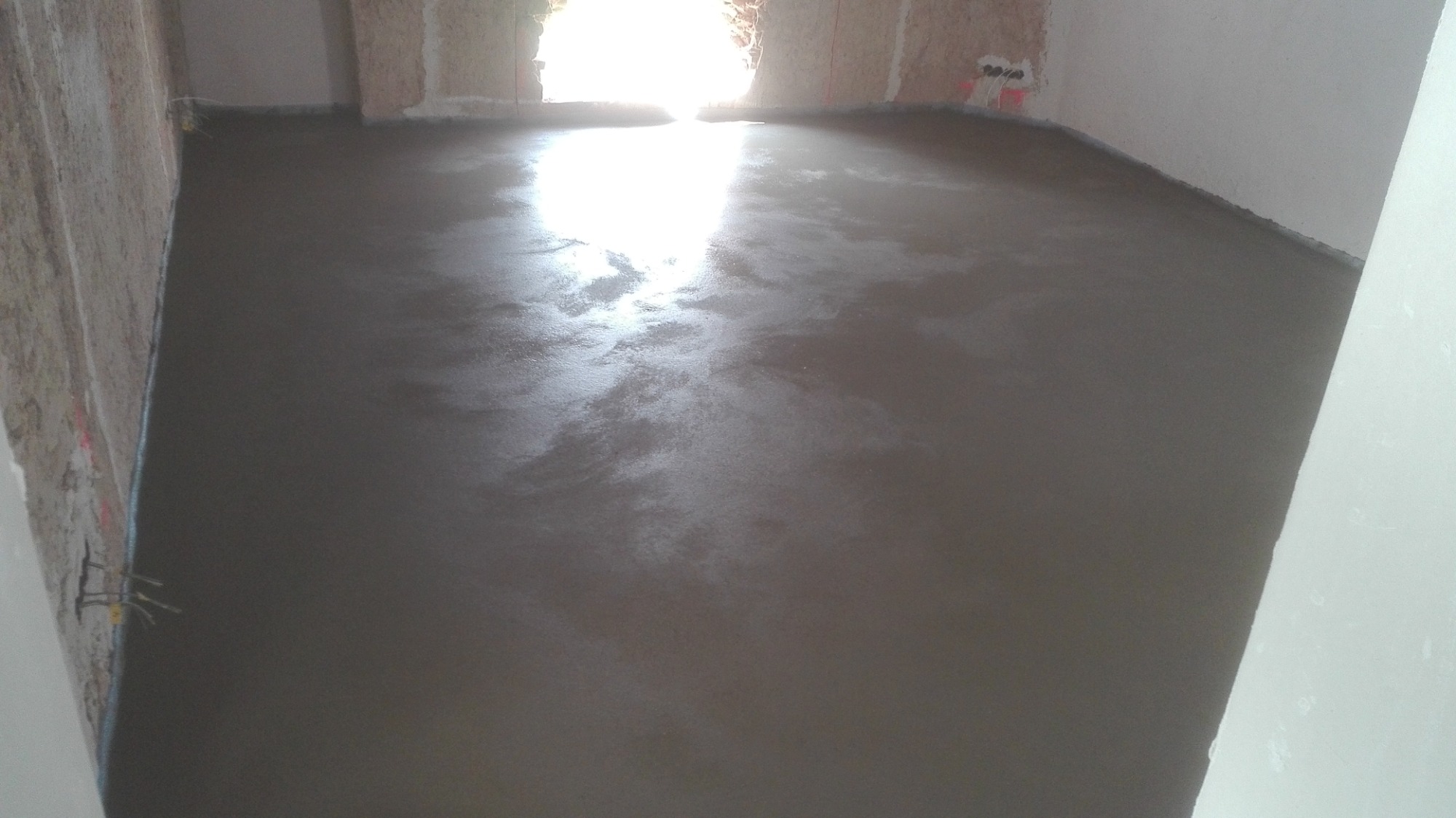 Świeżo wylana, mokra wylewka podłogowa w surowym pomieszczeniu z widocznymi fragmentami ścian w stanie deweloperskim, oświetlona przez jasne światło dzienne wpadające przez otwór drzwiowy.