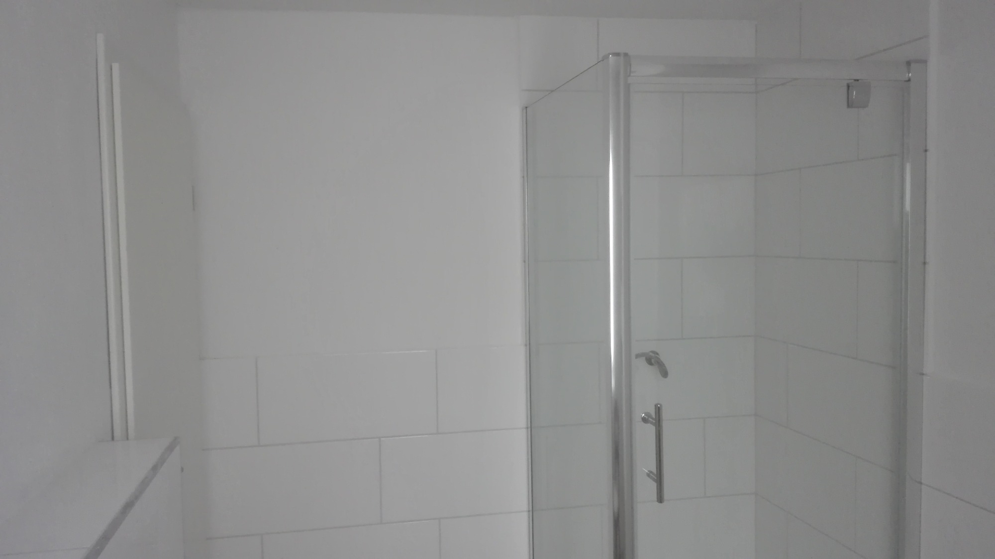 Kabina prysznicowa z chromowanymi okuciami i uchwytem, białe prostokątne płytki na ścianach, minimalistyczne wnętrze łazienki.