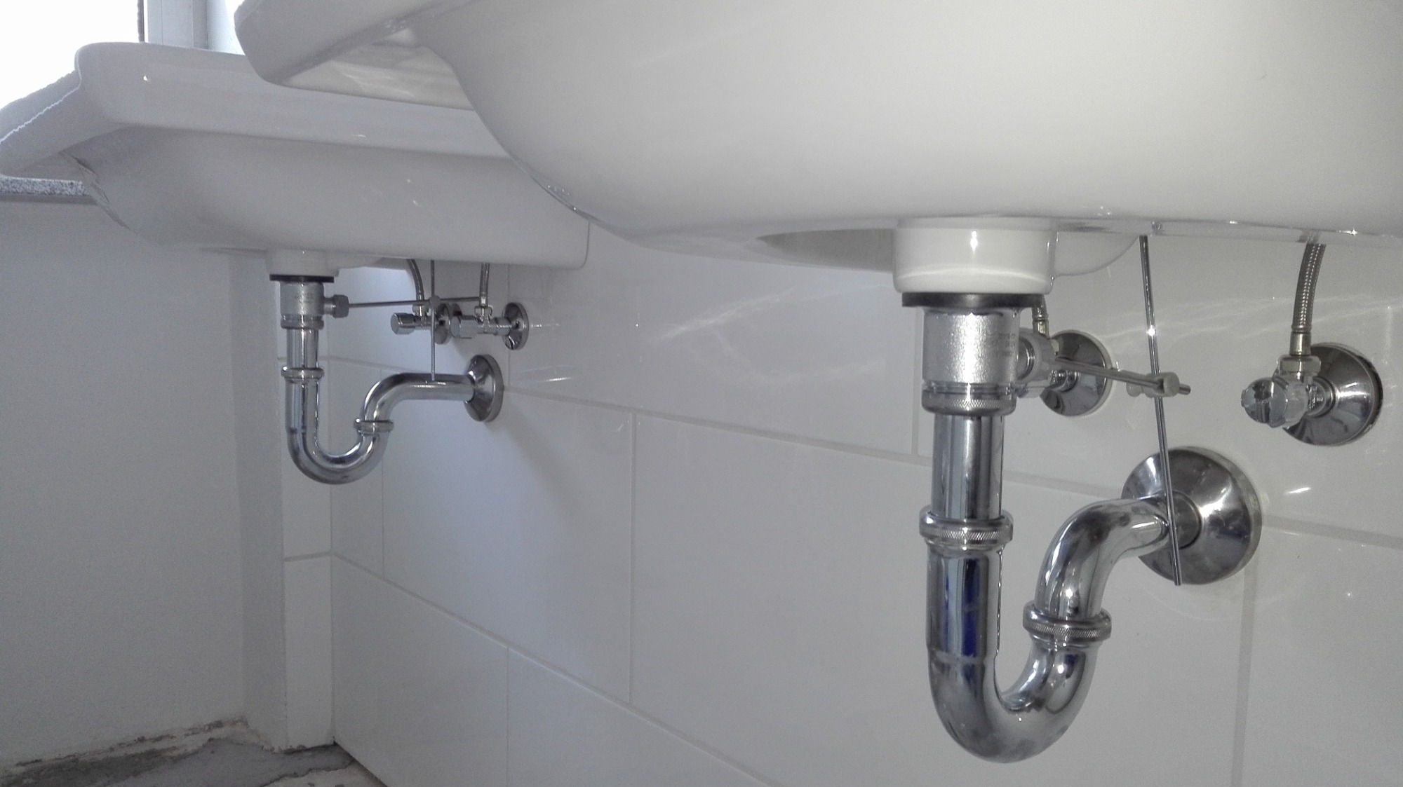 Dwie białe umywalki podwieszane z widocznymi chromowanymi syfonami i zaworami, zamontowane na ścianie wyłożonej białymi, prostokątnymi płytkami.