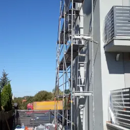 Dom jednorodzinny w trakcie ocieplania, widoczne rusztowania, otynkowana ściana, przygotowane materiały budowlane i młode tuje na pierwszym planie.