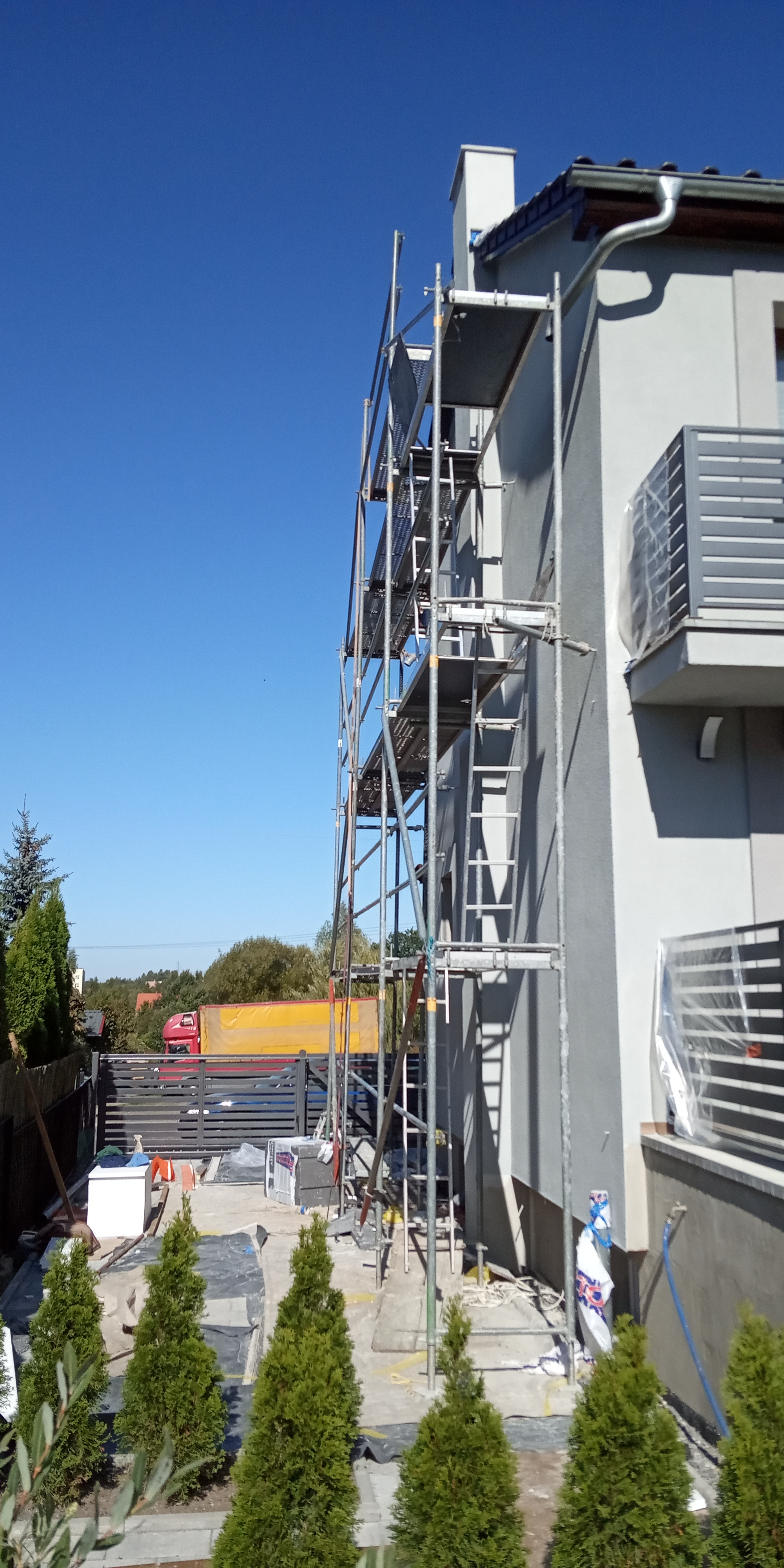 Dom jednorodzinny w trakcie ocieplania, widoczne rusztowania, otynkowana ściana, przygotowane materiały budowlane i młode tuje na pierwszym planie.