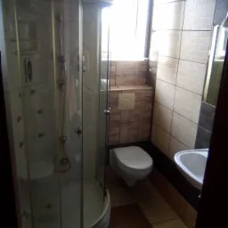 Kabina prysznicowa z hydromasażem, toaleta pod oknem i umywalka w łazience wykończonej brązowo-beżowymi płytkami.