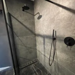 Nowoczesna kabina prysznicowa z czarną armaturą i szarymi płytkami imitującymi kamień, widoczny odpływ liniowy.