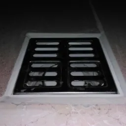 Kwadratowa, czarna kratka odpływowa w podłodze wyłożonej płytkami, uszczelniona białym silikonem.