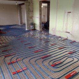 INSTAL-KRAK - Niedokończony montaż ogrzewania podłogowego w surowym stanie, widoczne nieotynkowane ściany, niebieskie rury na czerwonej siatce, narzędzia w tle, instalator w rogu pokoju.