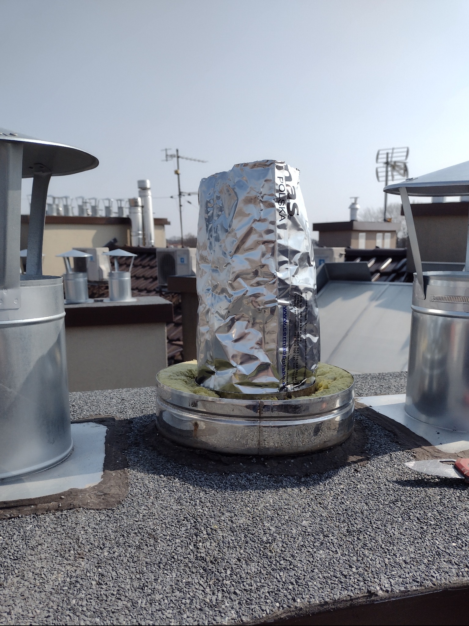 Orurowanie komina z izolacją owinięte folią aluminiową na dachu pokrytym żwirem, widoczne inne kominy w tle.