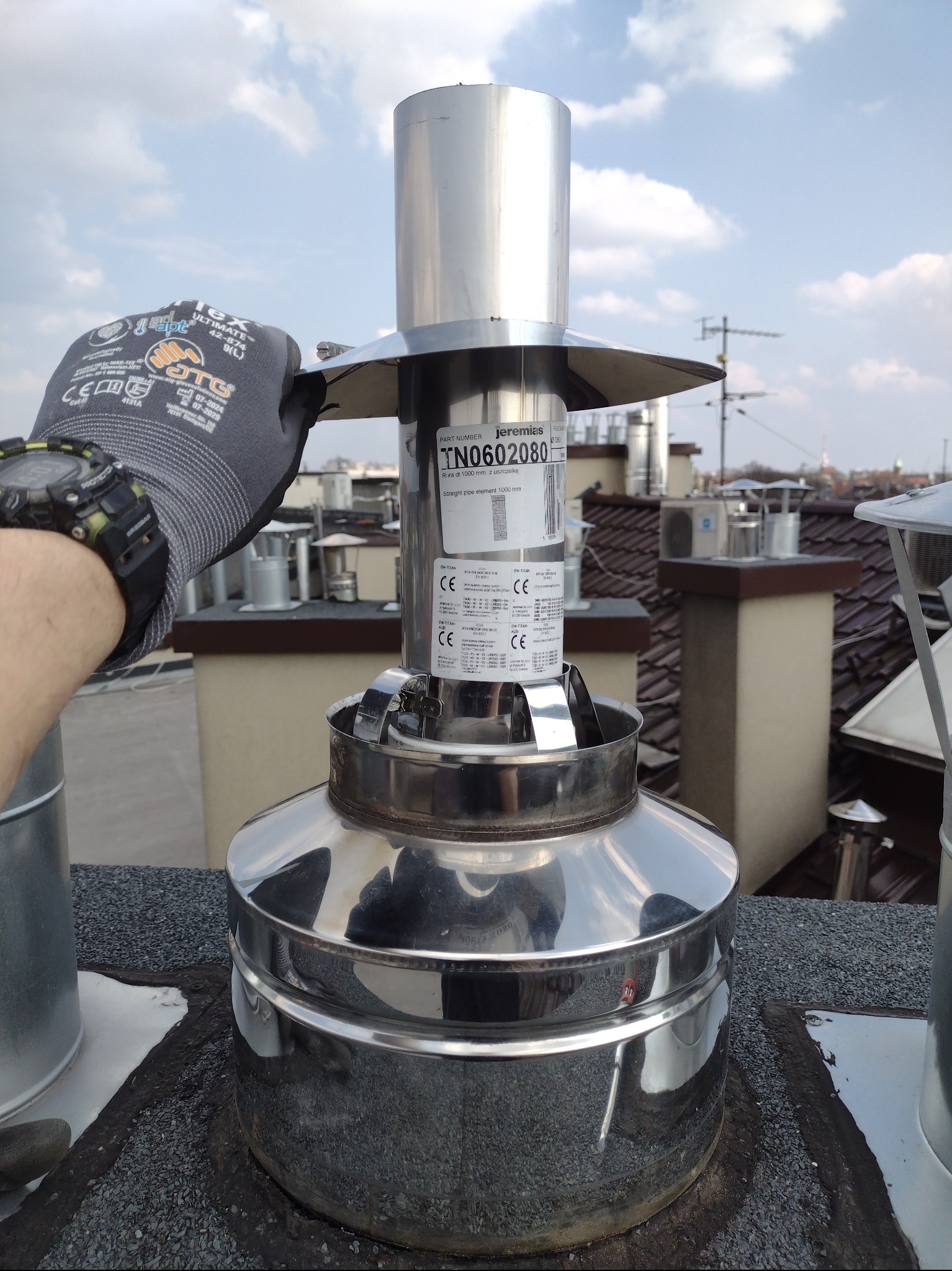 Montaż elementu kominowego Jeremias TN0602080 na dachu, widoczna ręka w rękawicy roboczej z zegarkiem, inne kominy w tle.