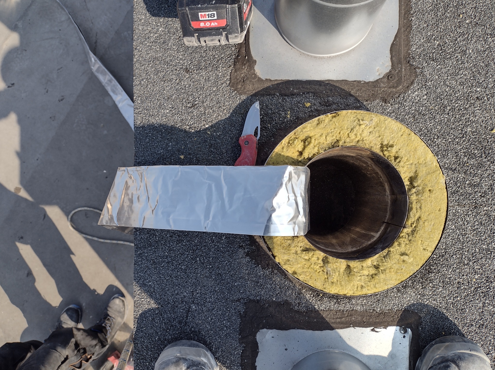 Otwór w dachu z izolacją, nożyk z czerwoną rączką, bateria do elektronarzędzi i taśma aluminiowa, widok z góry na etapie prac montażowych.