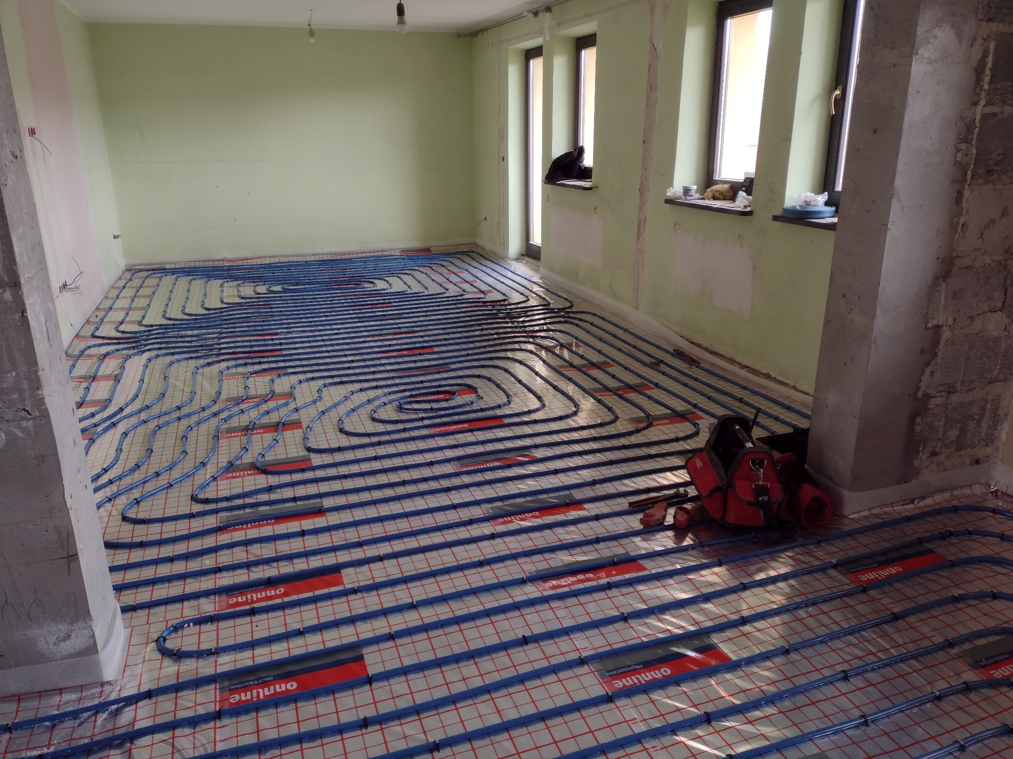 Niebieskie rury ogrzewania podłogowego ułożone na siatce w surowym pomieszczeniu z oknami i kolumną, widoczna czerwona torba narzędziowa.