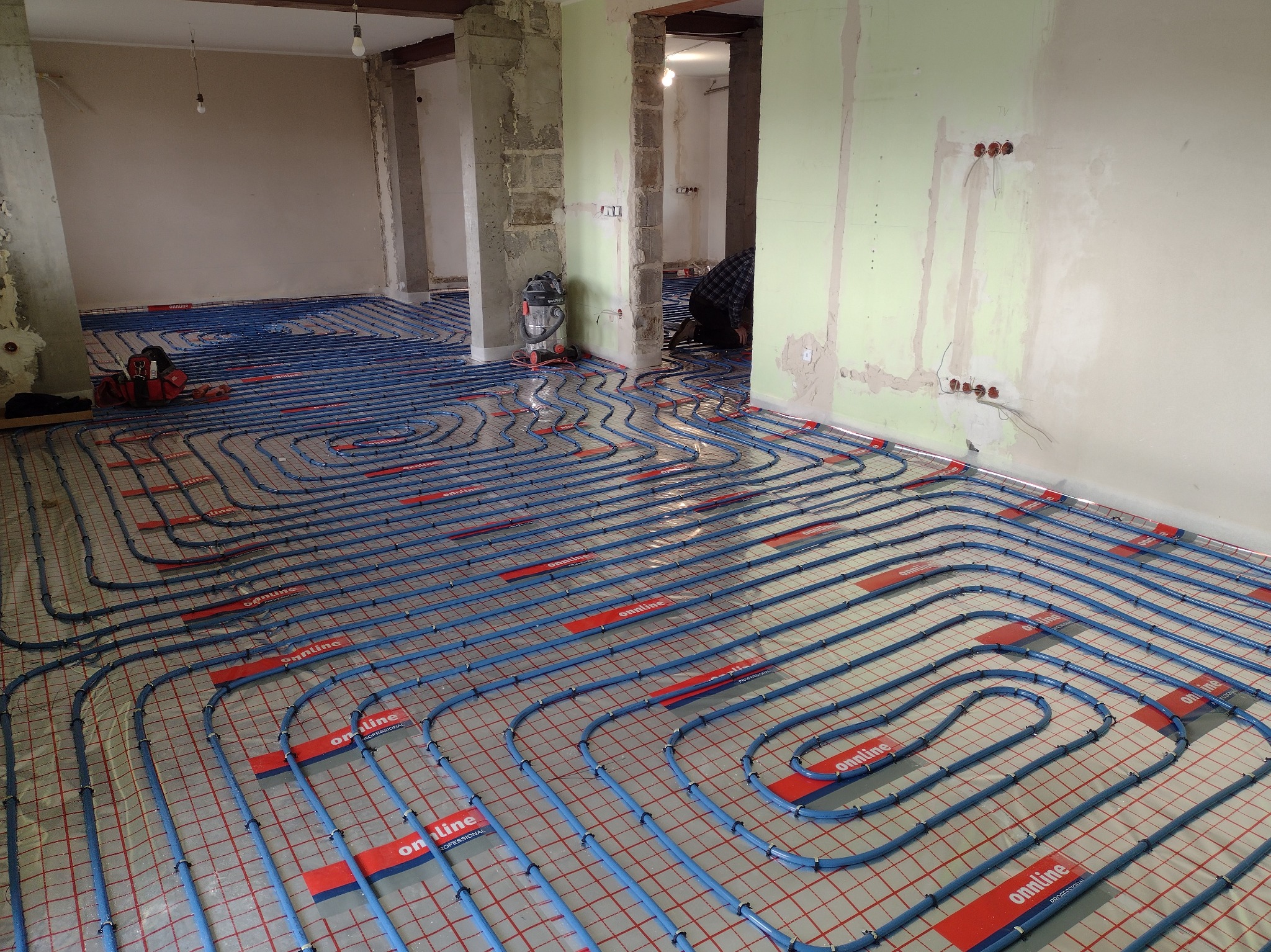 Niedokończony montaż ogrzewania podłogowego w surowym stanie, widoczne nieotynkowane ściany, niebieskie rury na czerwonej siatce, narzędzia w tle, instalator w rogu pokoju.